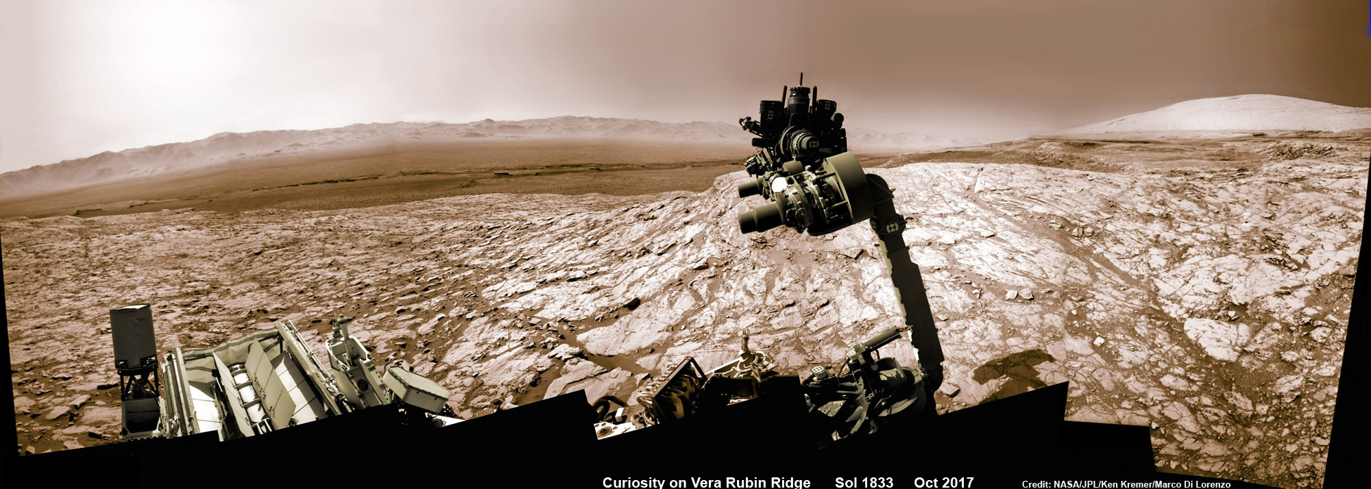  Марсоход НАСА Curiosity поднял роботизированную руку с буром, направленным в небо, во время исследования хребта Веры Рубин у основания горы Шарп в кратере Гейл — на фоне далёкого кратера. Эта мозаика, сделанная камерой navcam, была сшита из необработанных изображений, полученных во время сол 1833, 2 октября 2017 года, и раскрашена.