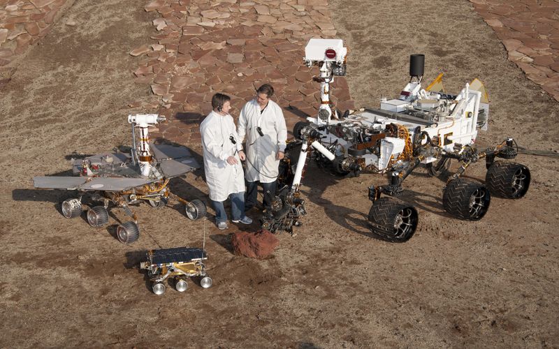 Photo of two people in white lab coats standing in a dry landscape surrounded by several wheeled robots.