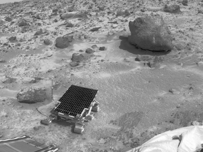 Black and white photo of a small wheeled robot on sandy terrain with a large rock in the background.