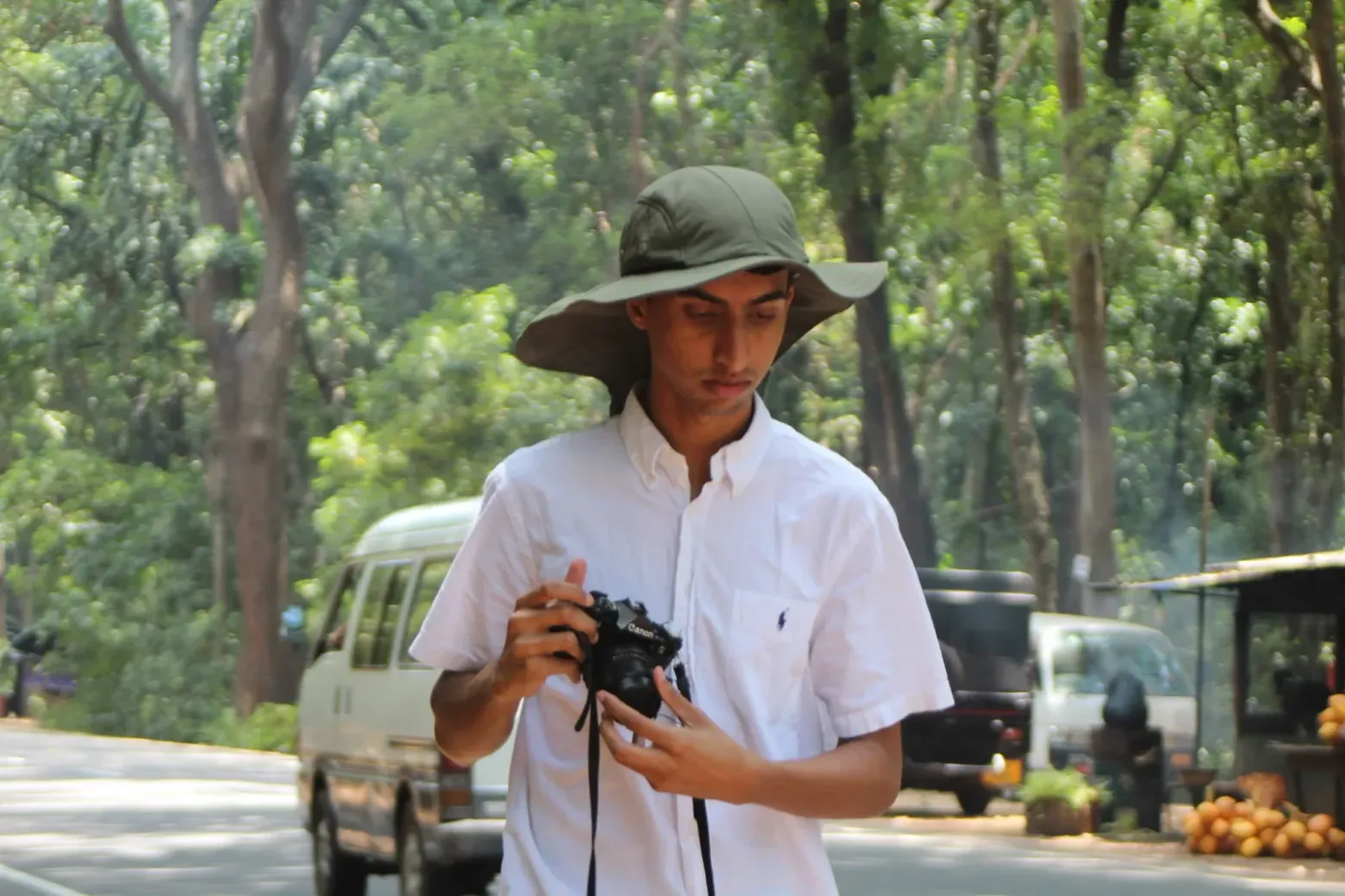 Shan in a wide-brimmed green hat and a white shirt, looking down at a black Canon film camera held in both hands, on a tree-lined road with a van and a stall of king coconuts behind him.