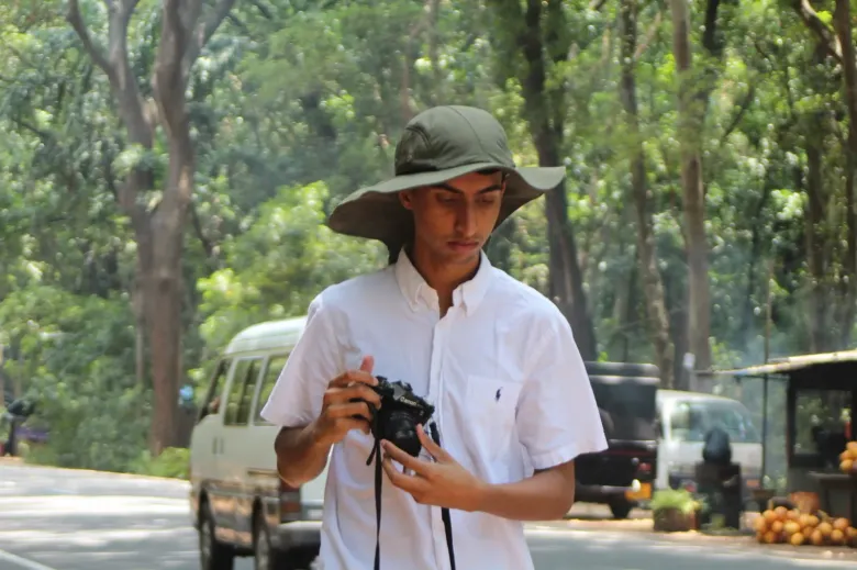Shan in a wide-brimmed green hat and a white shirt, looking down at a black Canon film camera held in both hands, on a tree-lined road with a van and a stall of king coconuts behind him.